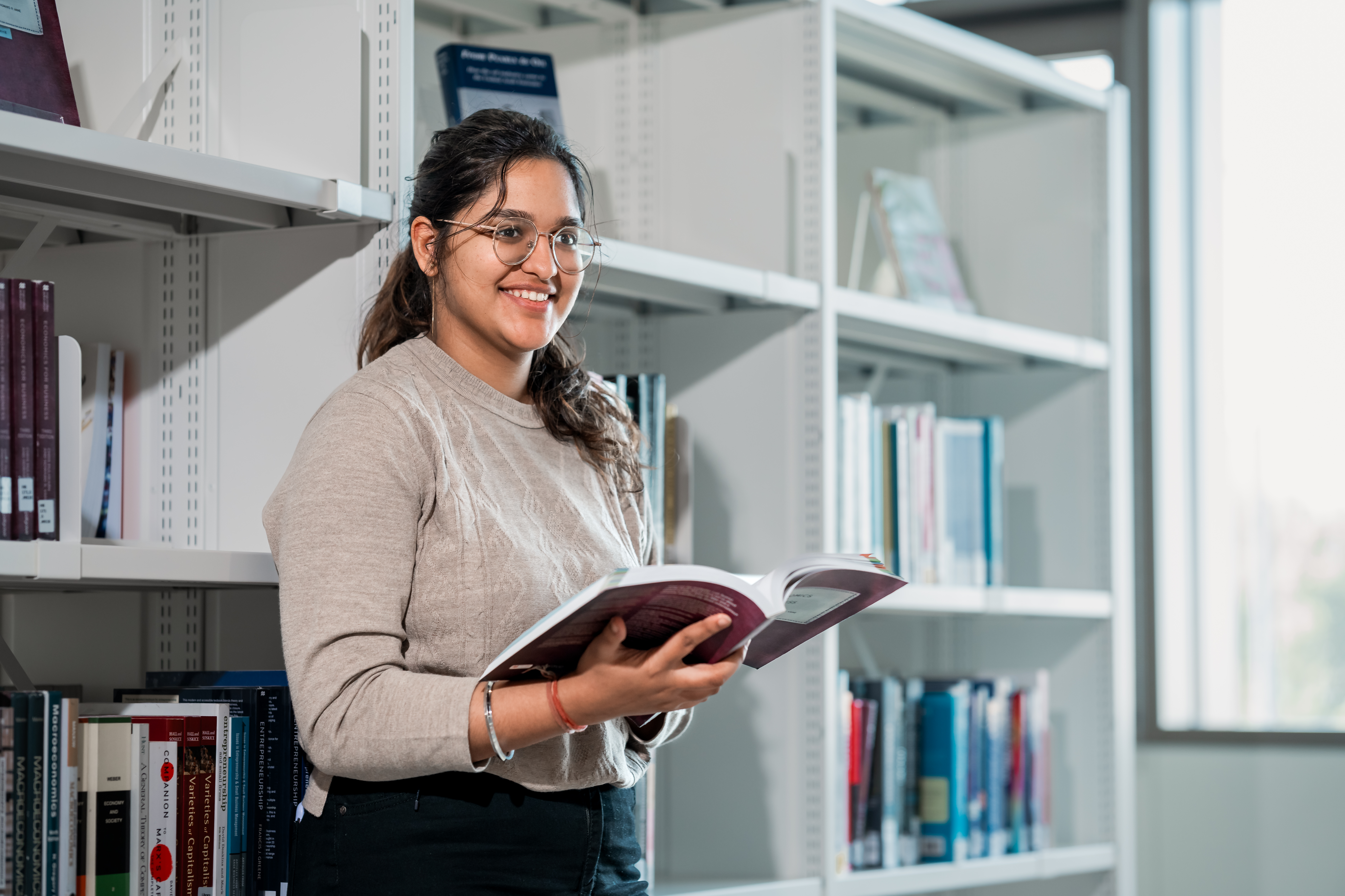 A student reads an open book standing against a bookshelf