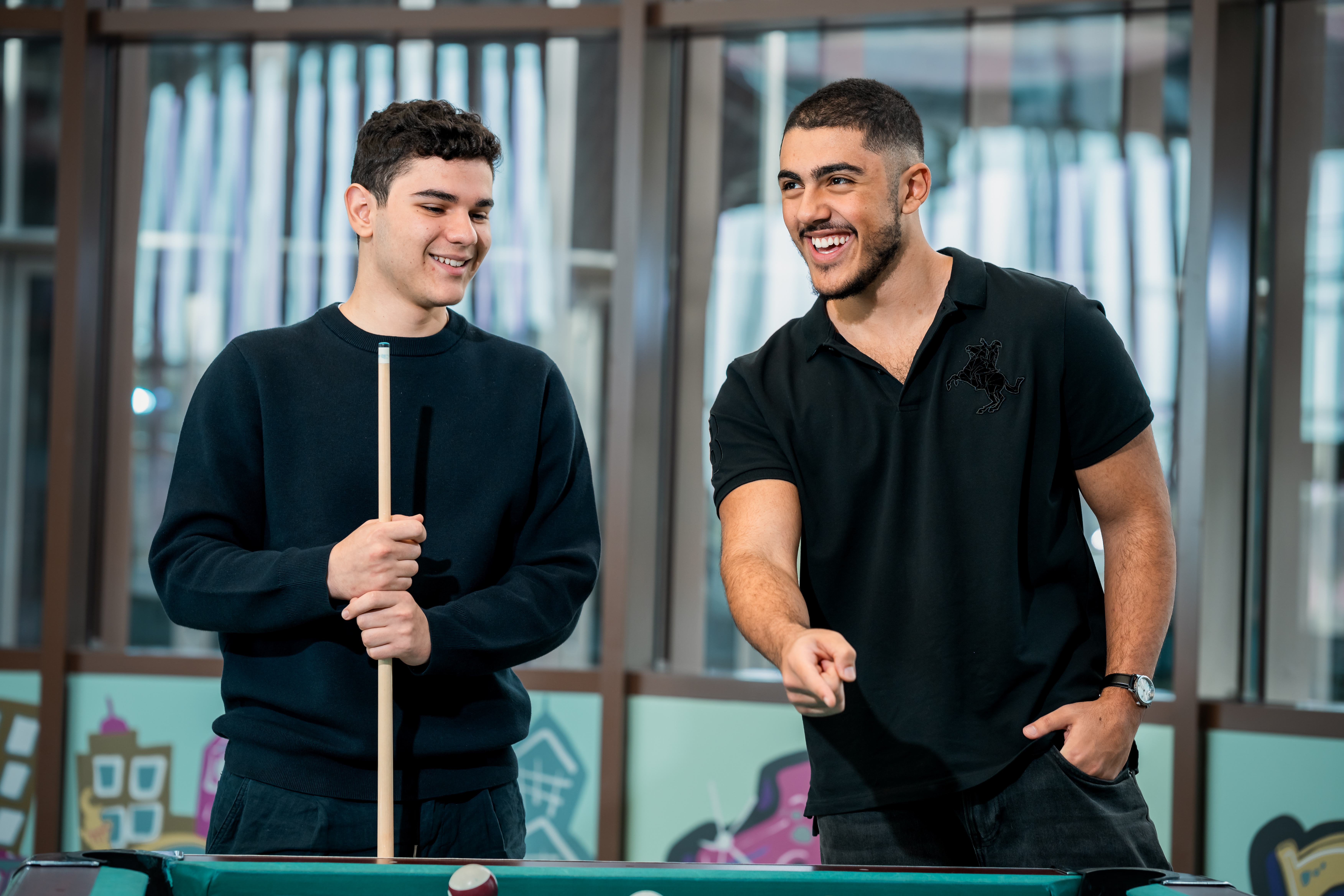Two students stand at a pool table, one holding a cue
