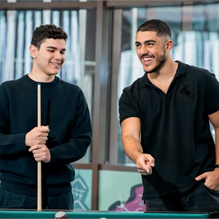 Two students stand at a pool table, one holding a cue